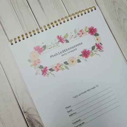 Floral spiral daily notepad with #behindthesuccess cover and contact info section on wood table