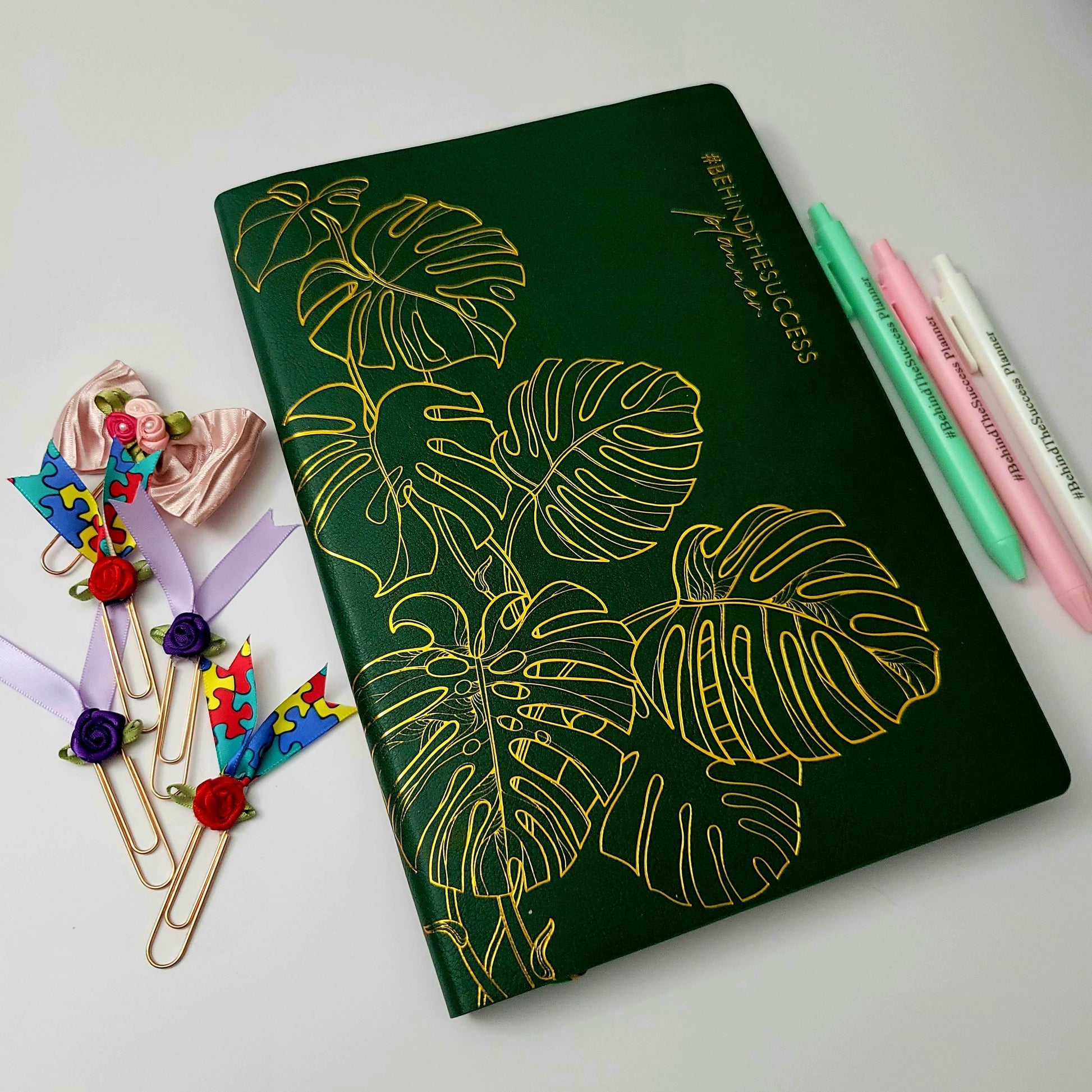 Green undated productivity planner with gold leaf design, colorful pens, and floral paper clips