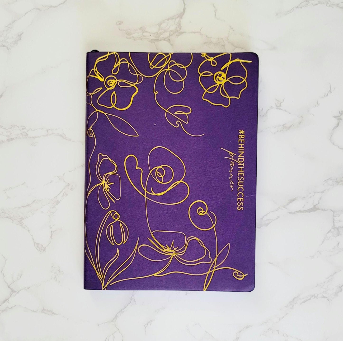 Purple productivity planner with yellow floral design and #BEHINDTHESUCCESS on marble surface