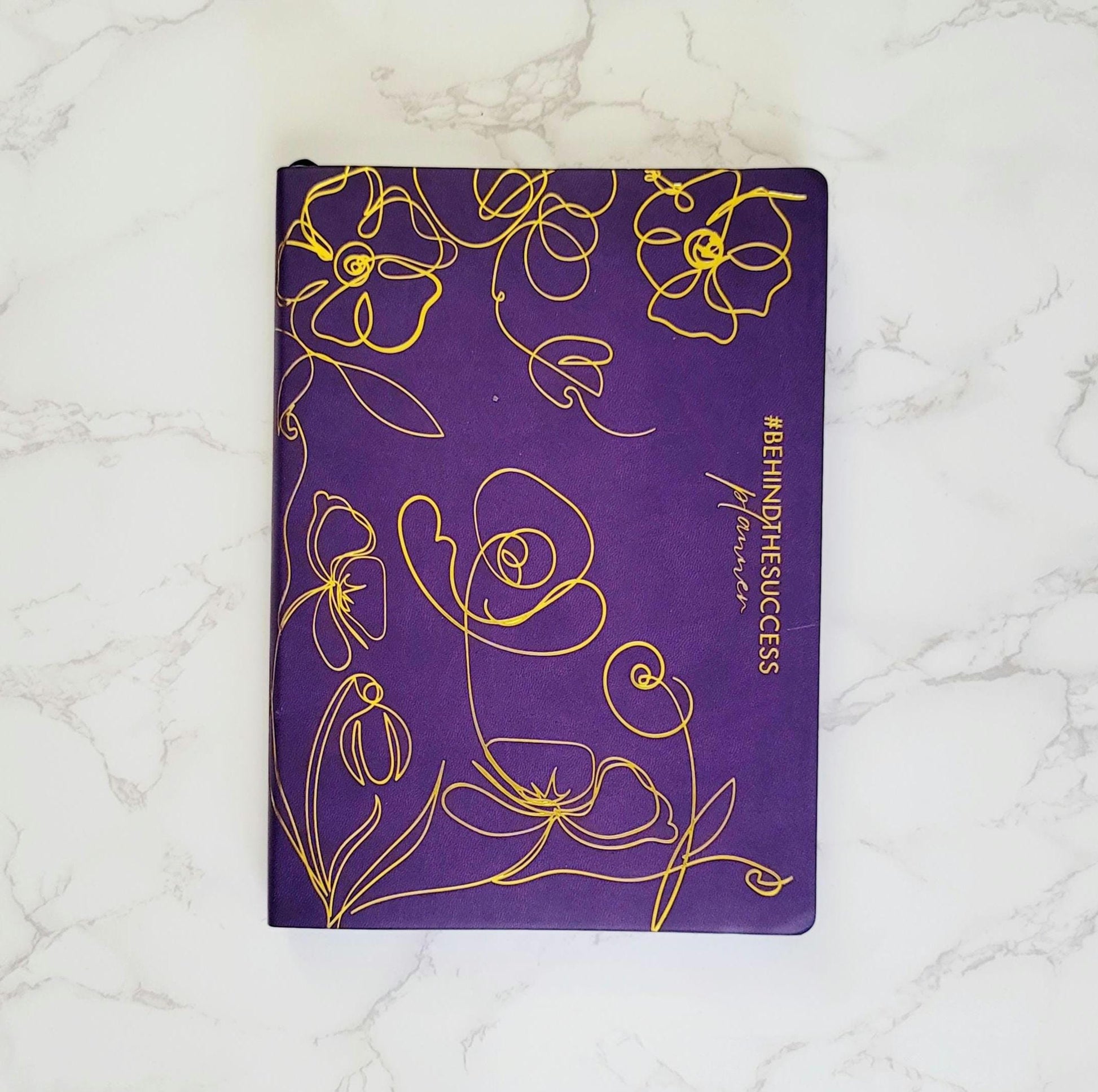 Purple productivity planner with yellow floral design and #BEHINDTHESUCCESS on marble surface