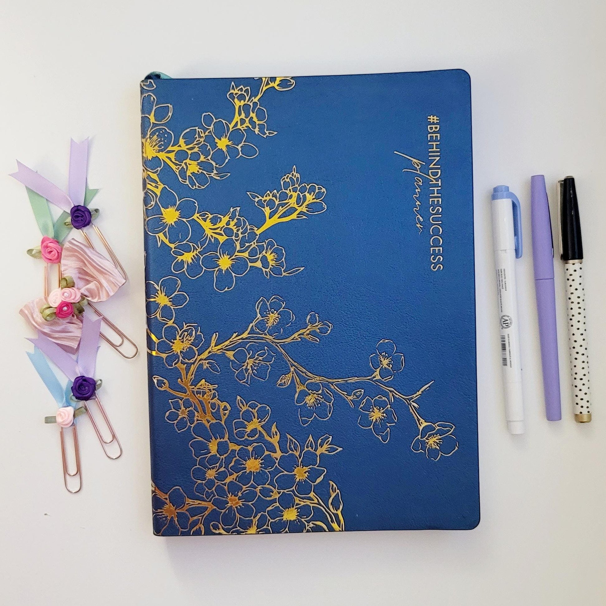 Blue floral undated planner with gold accents, decorative paper clips, and pens on a white desk