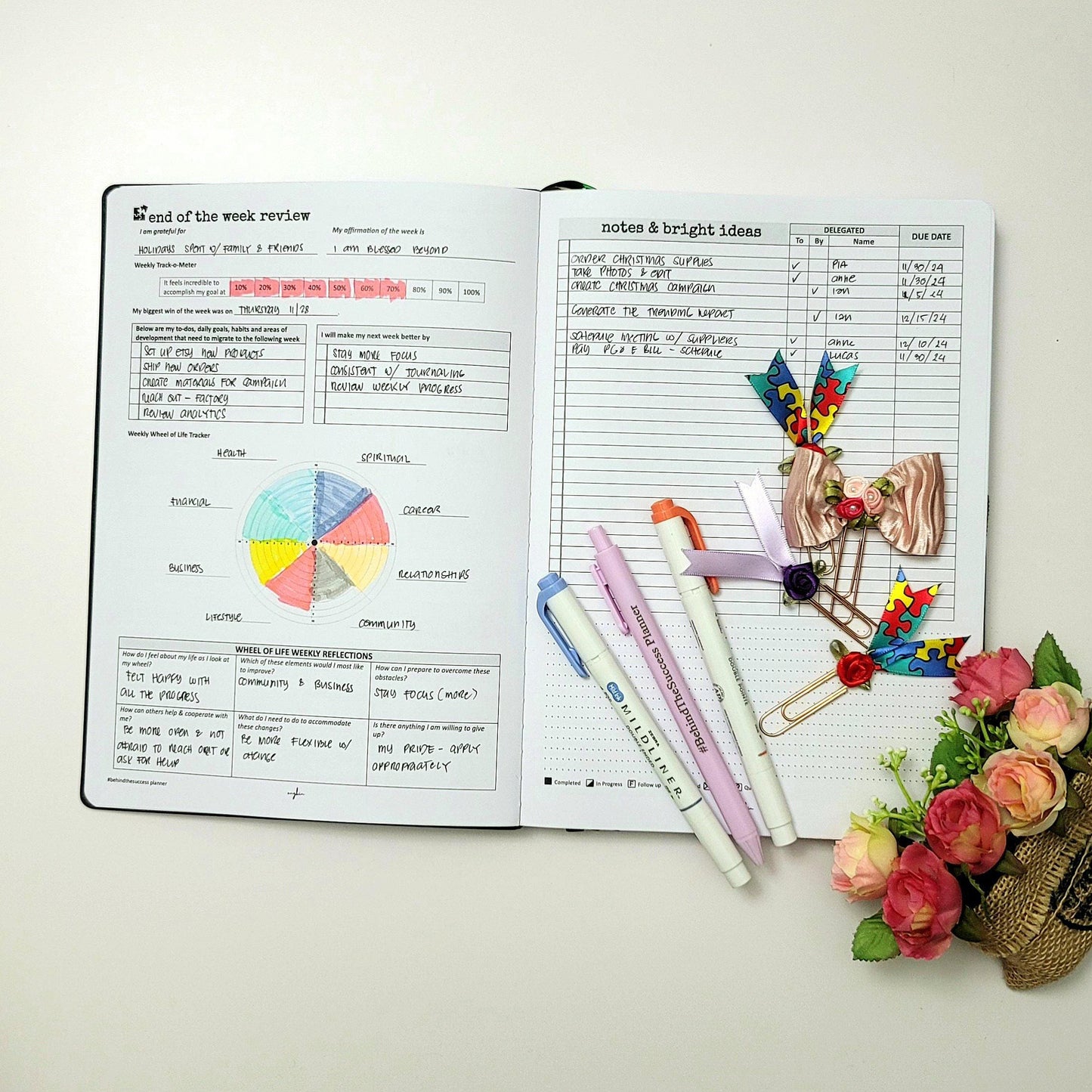 Open productivity life planner with colorful pens, paper clips, and flowers on white desk