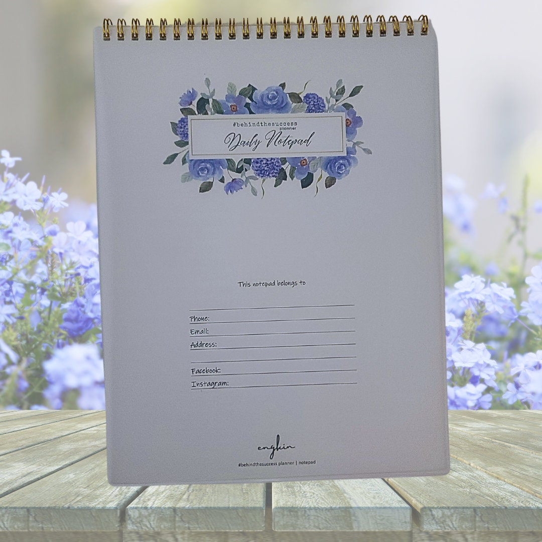 Spiral-bound daily notepad with floral cover, contact info section, and violet flowers in background