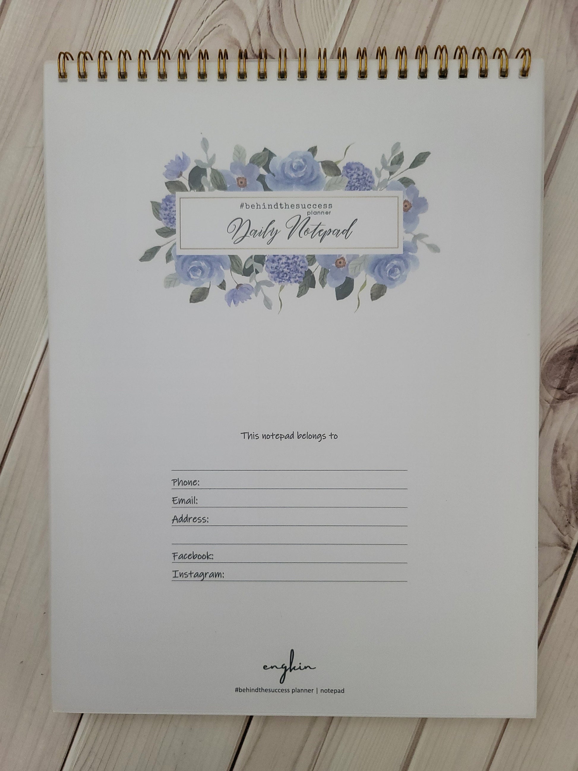 Spiral-bound daily notepad planner with blue floral design and contact info fields on white wood background.