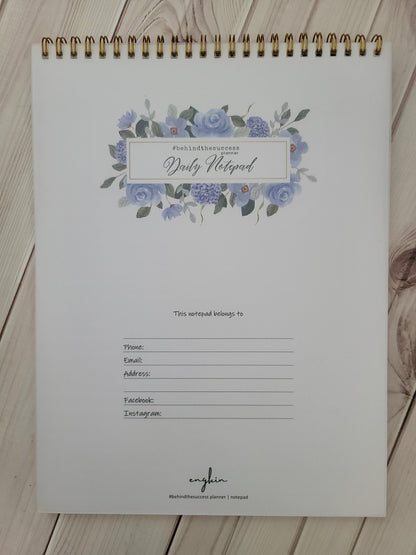 Spiral-bound daily notepad planner with blue floral design and contact info fields on white wood background.